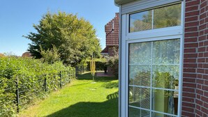A house with a large glass window, a brick wall, and a grassy yard with trees. - Vacation home in Nessmersiel 20-007a - 200-007a (Dornum)