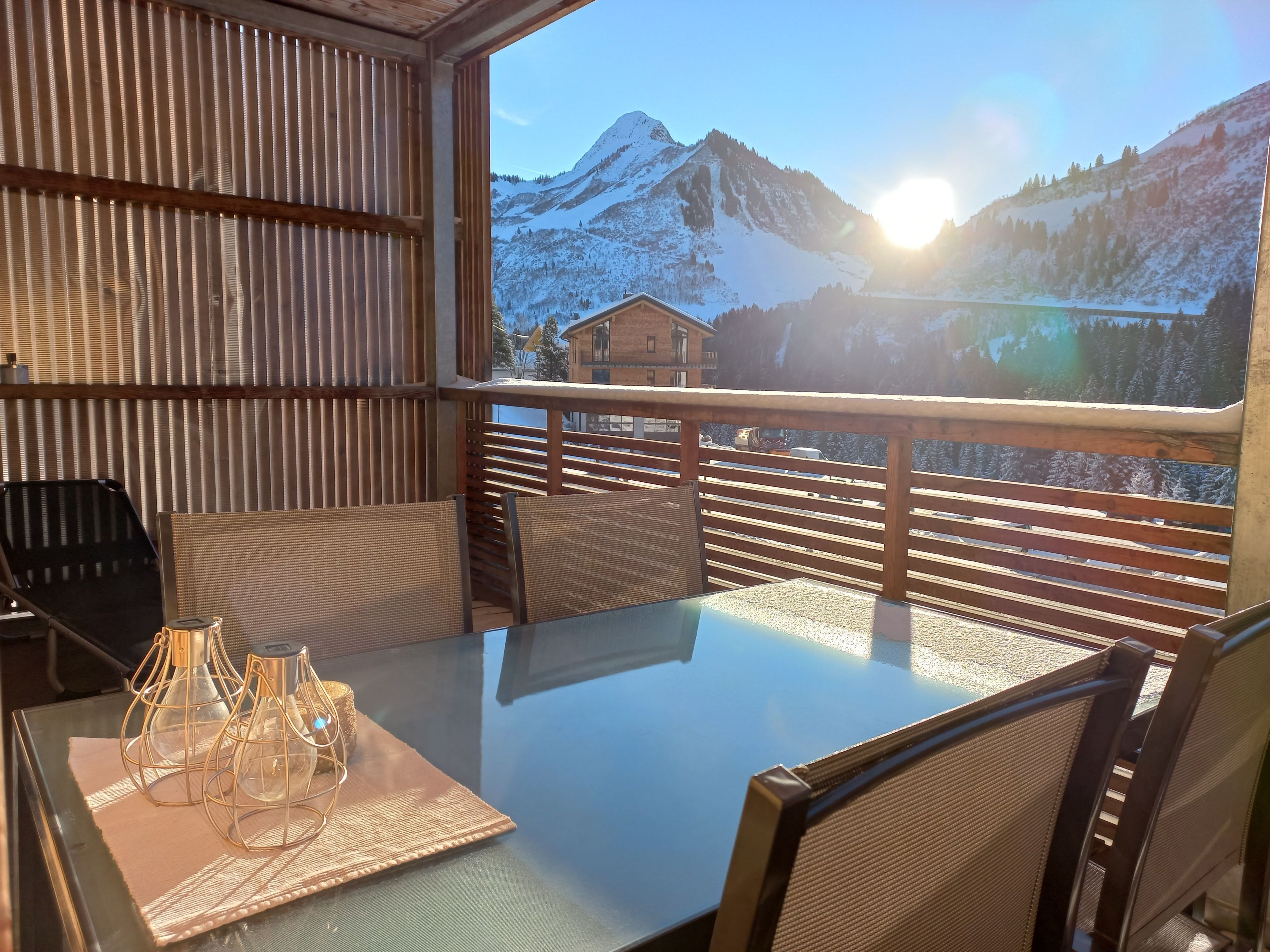 Un balcon avec une table et des chaises, une table à plateau en verre, et une vue sur des montagnes enneigées.