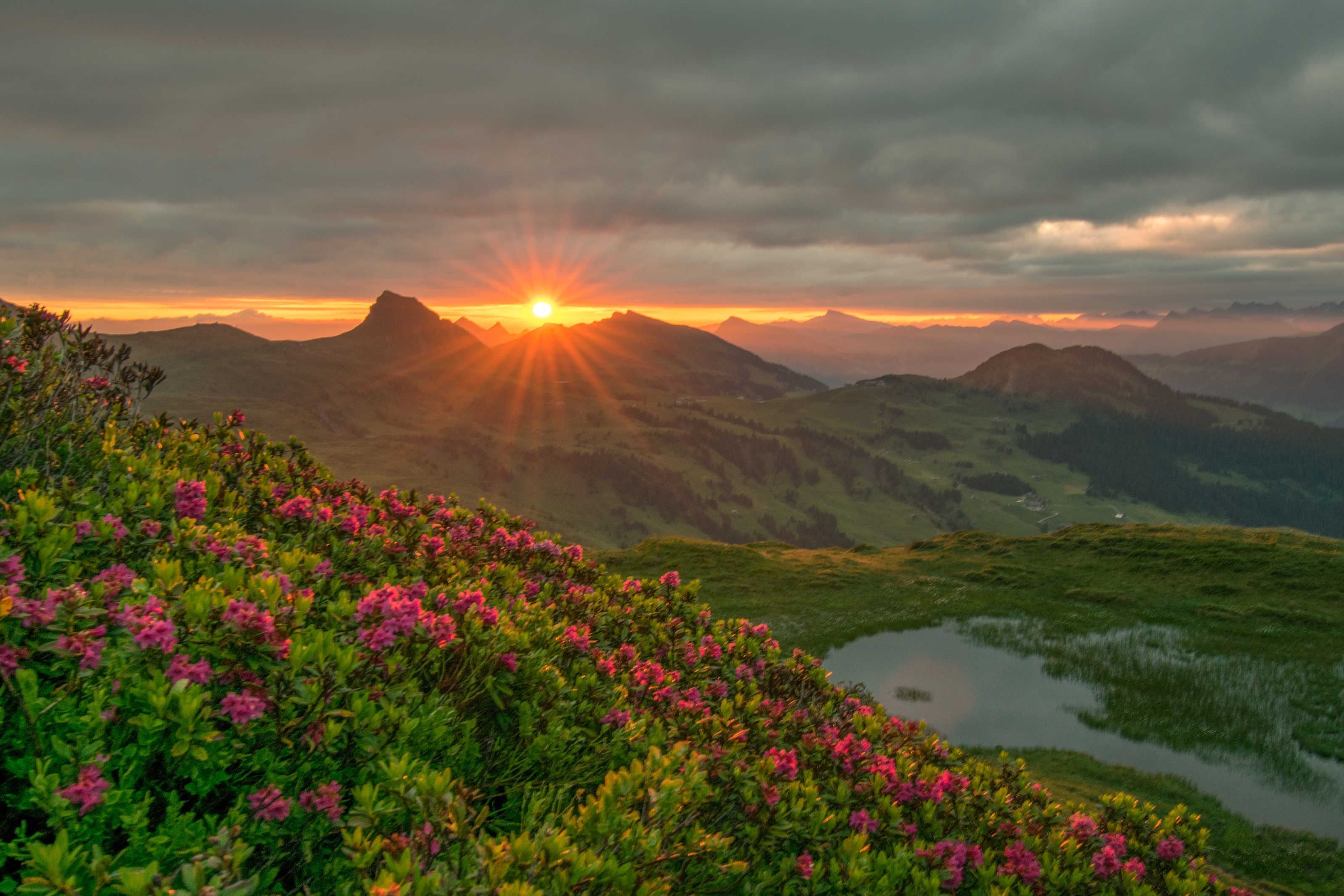 A mountainous landscape with a sunrise, green hills, and a body of water.