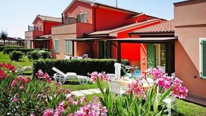 Un immeuble à plusieurs étages doté d’une façade rouge, d’un patio couvert et d’un jardin bien entretenu agrémenté de fleurs roses et de chaises blanches.