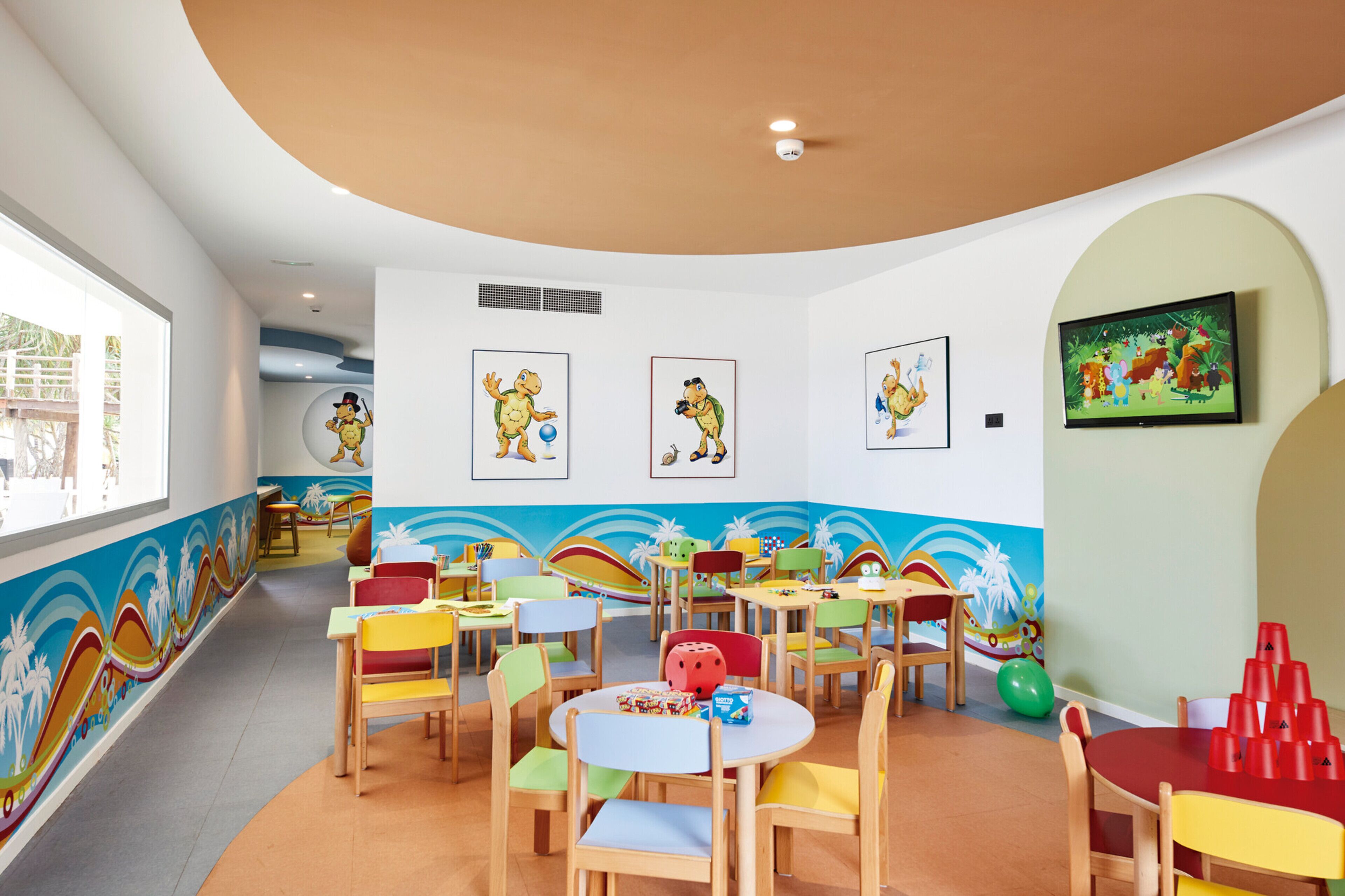 Zona de juegos para niños con sillas y mesas coloridas, un mural en la pared y una pantalla de televisión mostrando un programa animado.