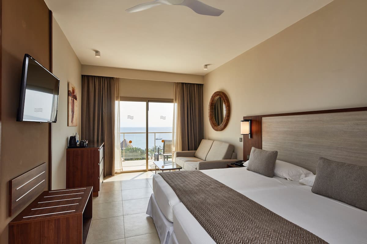 A hotel room with a large bed, a TV, a desk, a chair, a balcony with a view, and a ceiling fan.