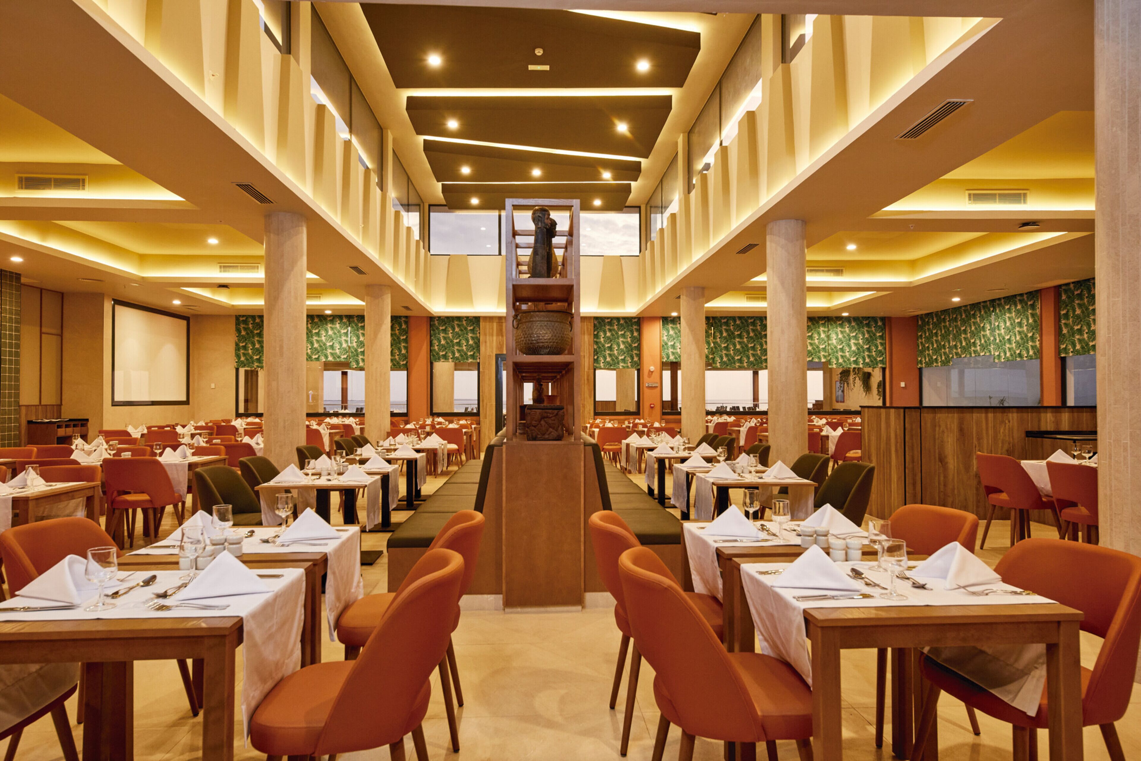 A spacious dining area with multiple tables and chairs, a central decorative column, and a large screen at the front.