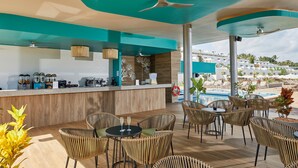 Un espace bar extérieur moderne, doté de mobilier en osier, offrant une vue sur la piscine et agrémenté d’un ventilateur de plafond.