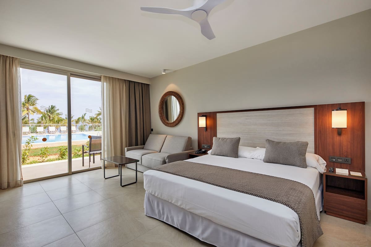A hotel room with a large bed, a desk, a chair, a nightstand, a lamp, and a view of a swimming pool.