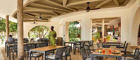 An outdoor dining area with wooden tables and chairs, a waiter in uniform, and a view of the restaurant's interior.