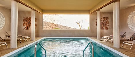 Una piscina interior con agua cristalina, rodeada de sillones de descanso y columnas, con vista a un paisaje rocoso a través de un ventanal grande.