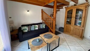 Una sala de estar con una escalera de madera, un sofá negro con cojines estampados y un aparador de madera con puertas de cristal.