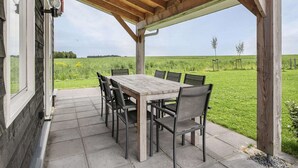 Une terrasse extérieure avec une table et des chaises en bois, un espace couvert doté d’un plafond en bois, et une vue sur un champ verdoyant.