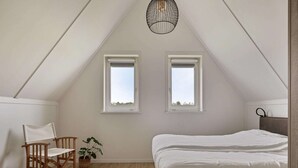 Une chambre avec un plafond en pente, deux fenêtres, un lit, une chaise et un luminaire suspendu.