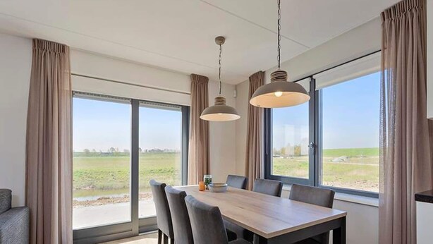Un espace de salle à manger moderne, doté d’une grande table et de chaises, de grandes fenêtres avec des rideaux et de luminaires suspendus.