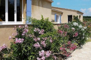 A house with a beige exterior, blue shutters, and a garden with pink flowers.