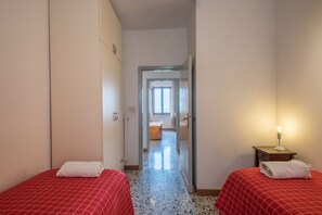 Ein Hotelzimmer mit zwei Betten, einem zur anderen Kammer führenden Flur und einem Nachttisch mit Lampe.