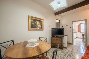 Ein Essbereich mit einem runden Holztisch, einer weißen Tischdecke, einem Gemälde an der Wand, einem Fernseher auf einem Holzständer und einer Küche im Hintergrund.