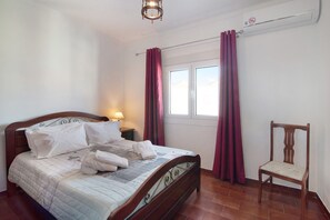 A neatly made bed with pillows and a blanket, a wooden chair, a nightstand with a lamp, and a window with curtains.