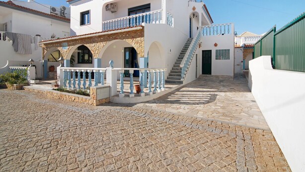 Un bâtiment blanc doté d’une cour pavée de pierres, d’un balcon avec des balustrades bleues et d’un escalier menant à l’entrée.