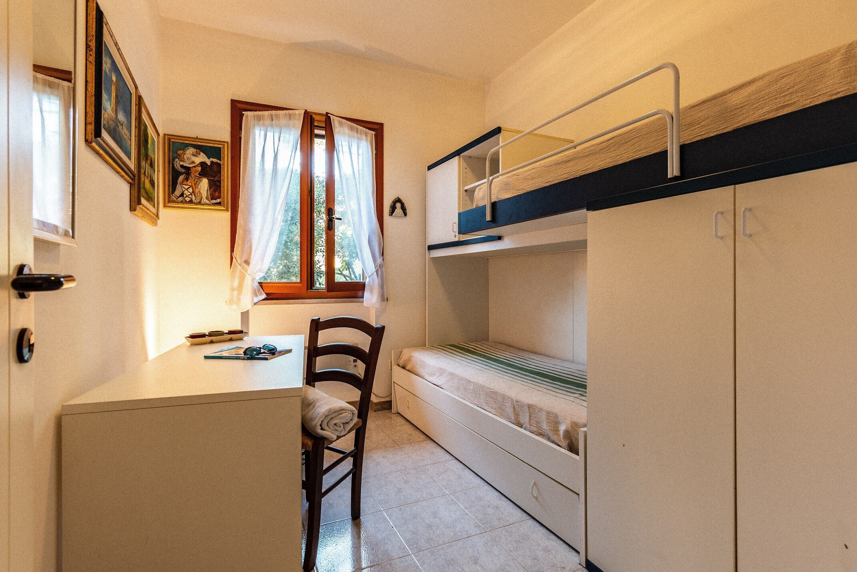Een kleine kamer met een stapelbed, een bureau, een stoel, een klein kastje en een raam met gordijnen.