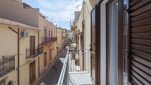 Una calle estrecha flanqueada por edificios, un balcón con barandilla metálica y una puerta con contraventanas de madera.