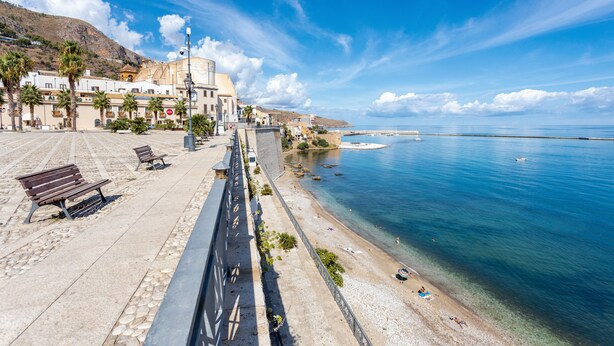 Un paseo marítimo con bancos, vista despejada del mar y edificios con balcones.
