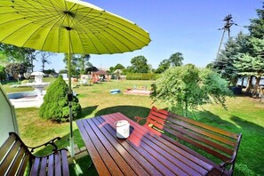 Een houten bank met een wit voorwerp, een gele parasol, en een tuin met een fontein en bomen.