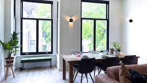 A dining area with a wooden table set for four, surrounded by chairs, with large windows offering a view of trees and buildings. - LVM Stays (The Hague)