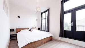 A bedroom with a wooden bed, a hanging light fixture, a nightstand with a lamp, and large windows with curtains. - LVM Stays (The Hague)