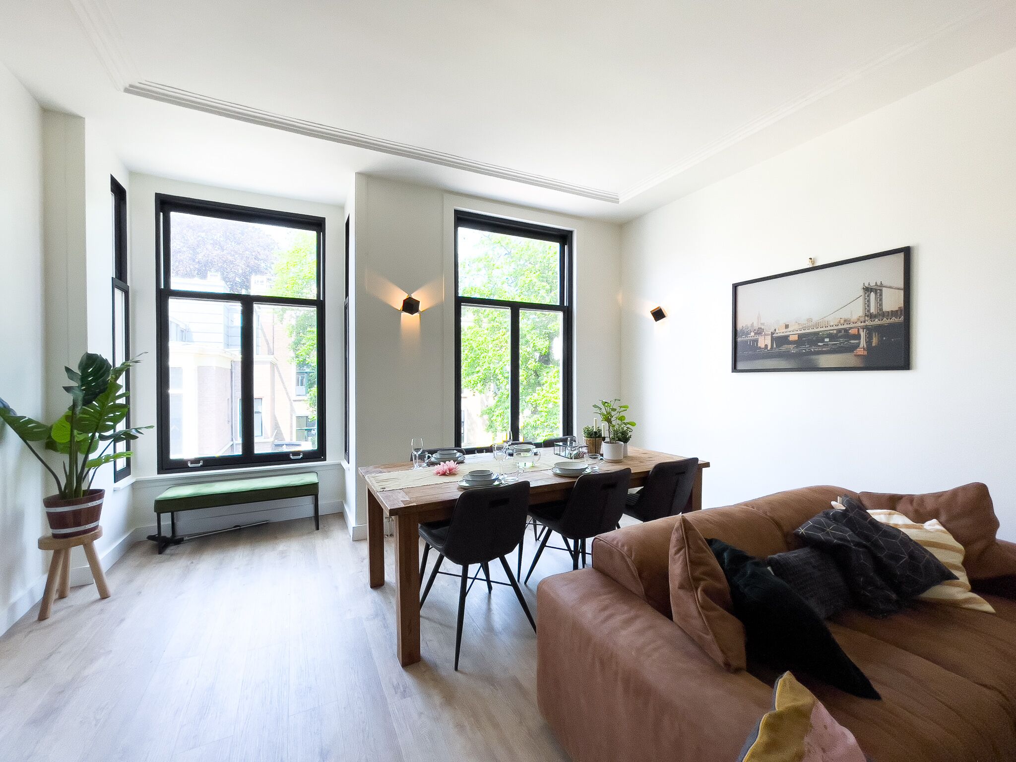 A dining area with a wooden table, chairs, and a bench. There are large windows with black frames, a potted plant, and a framed picture on the wall.