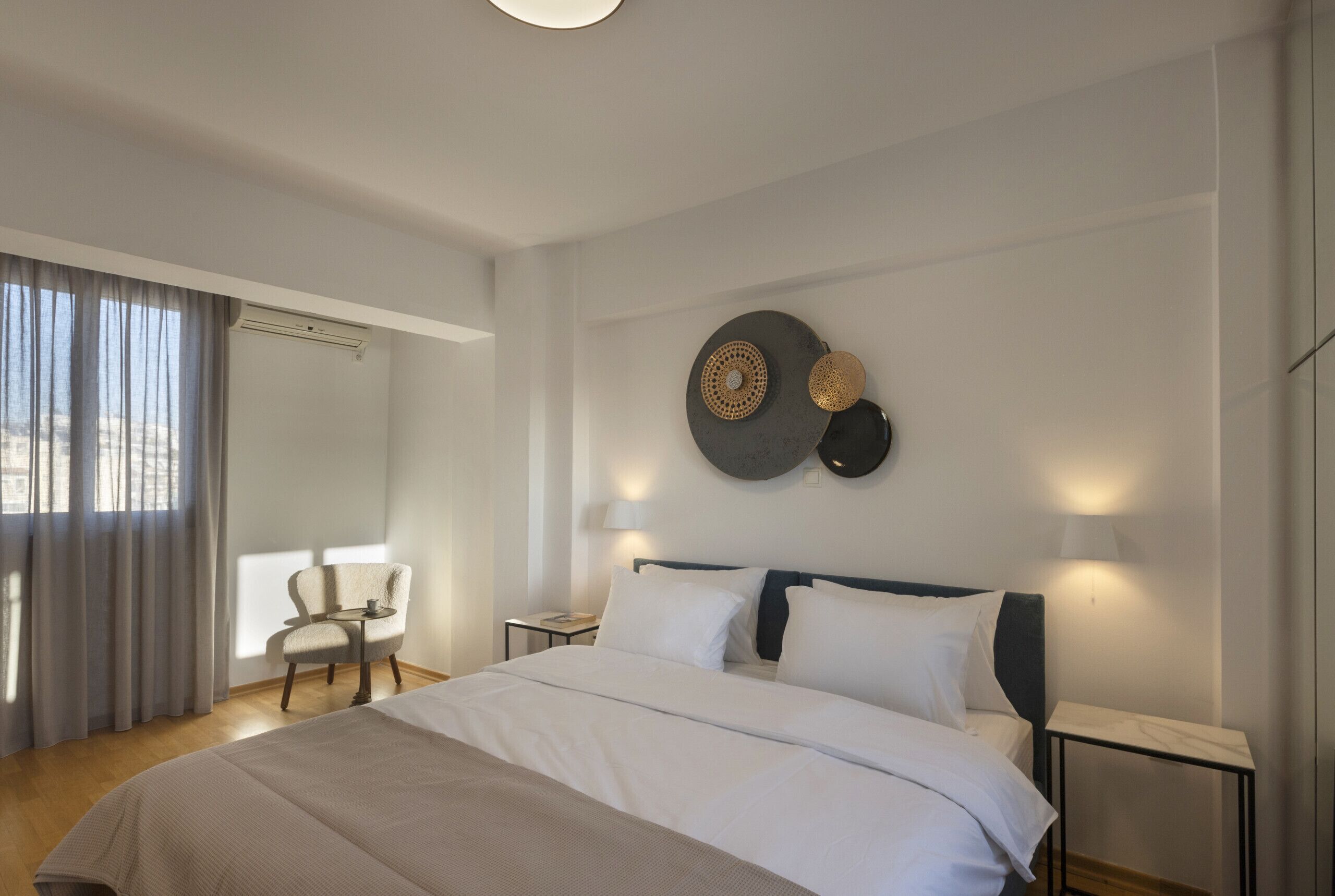 Ein modernes Hotelzimmer mit einem großen Bett, einem Sessel, einem Schreibtisch und Blick auf die Stadt durch das Fenster.