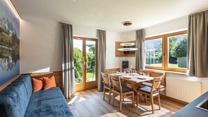 Un salon moderne avec un canapé bleu, une table à manger et des chaises, de grandes fenêtres avec des rideaux et une vue sur la verdure à l’extérieur.