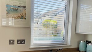 A kitchen with a built-in sink, a window with blinds, a framed picture of Newquay Harbour, and a teal-colored kettle.