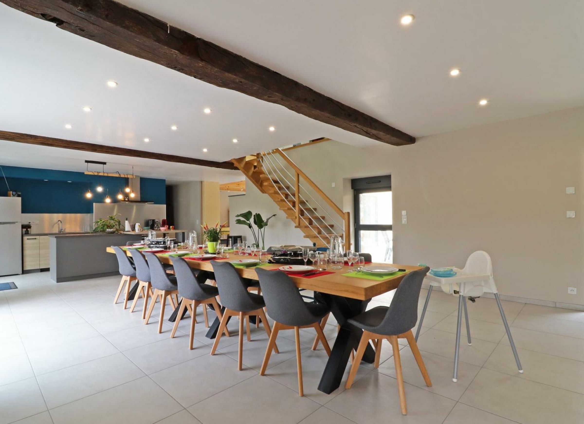 Un vaste espace dédié à la salle à manger, avec une longue table en bois et de nombreuses chaises, une cuisine à l’arrière-plan et un escalier menant à un niveau supérieur.