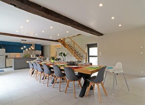 A spacious dining area with a long wooden table and multiple chairs, a kitchen in the background, and a staircase leading to an upper level.
