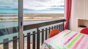 A balcony with a view of the sea and beach, a table with a striped tablecloth, and a red chair.