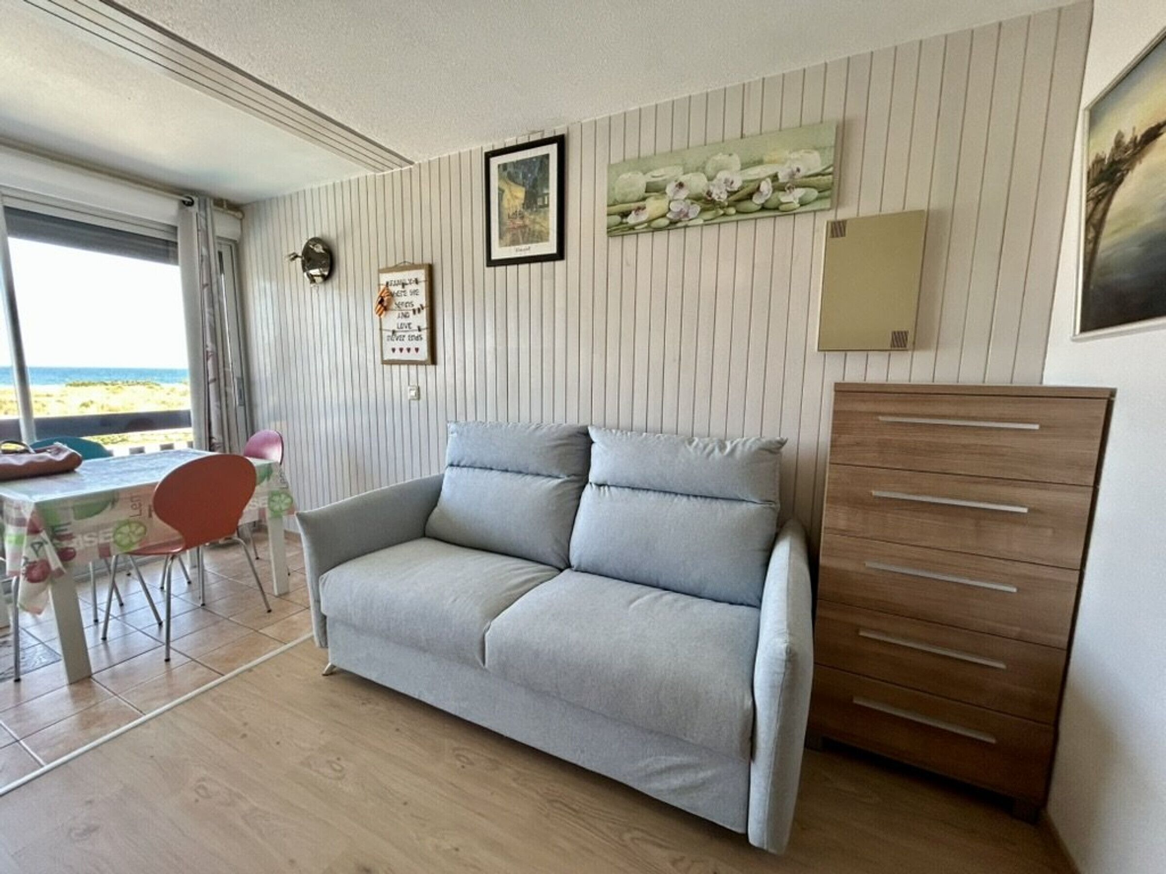 Une pièce avec un canapé gris, une commode en bois, une table à manger avec des chaises, et une vue sur la plage.