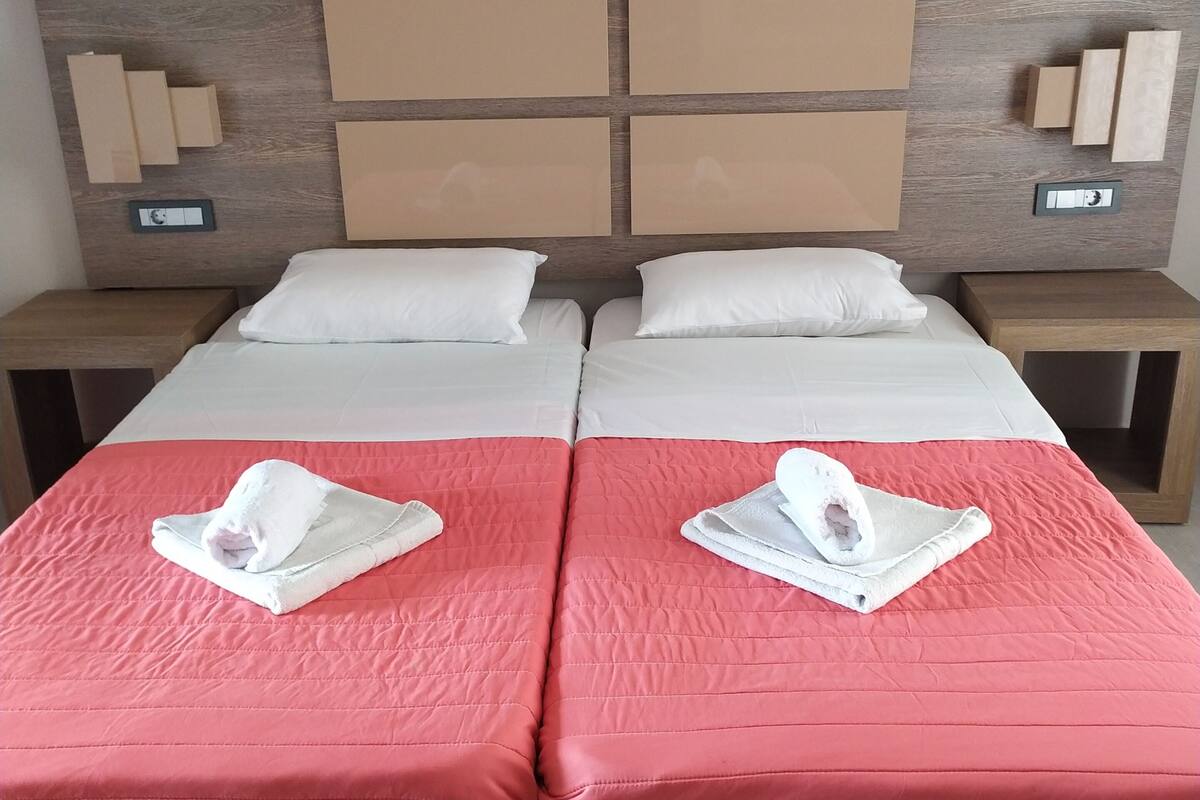 A double bed with two white pillows and a red bedspread. There are two nightstands with clocks and a wooden headboard with a light wood finish.