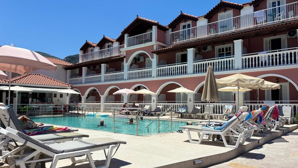 A pool area with lounge chairs and umbrellas, a building with balconies, and a clear blue sky. - Giannis Studios Alykes (Zakynthos)