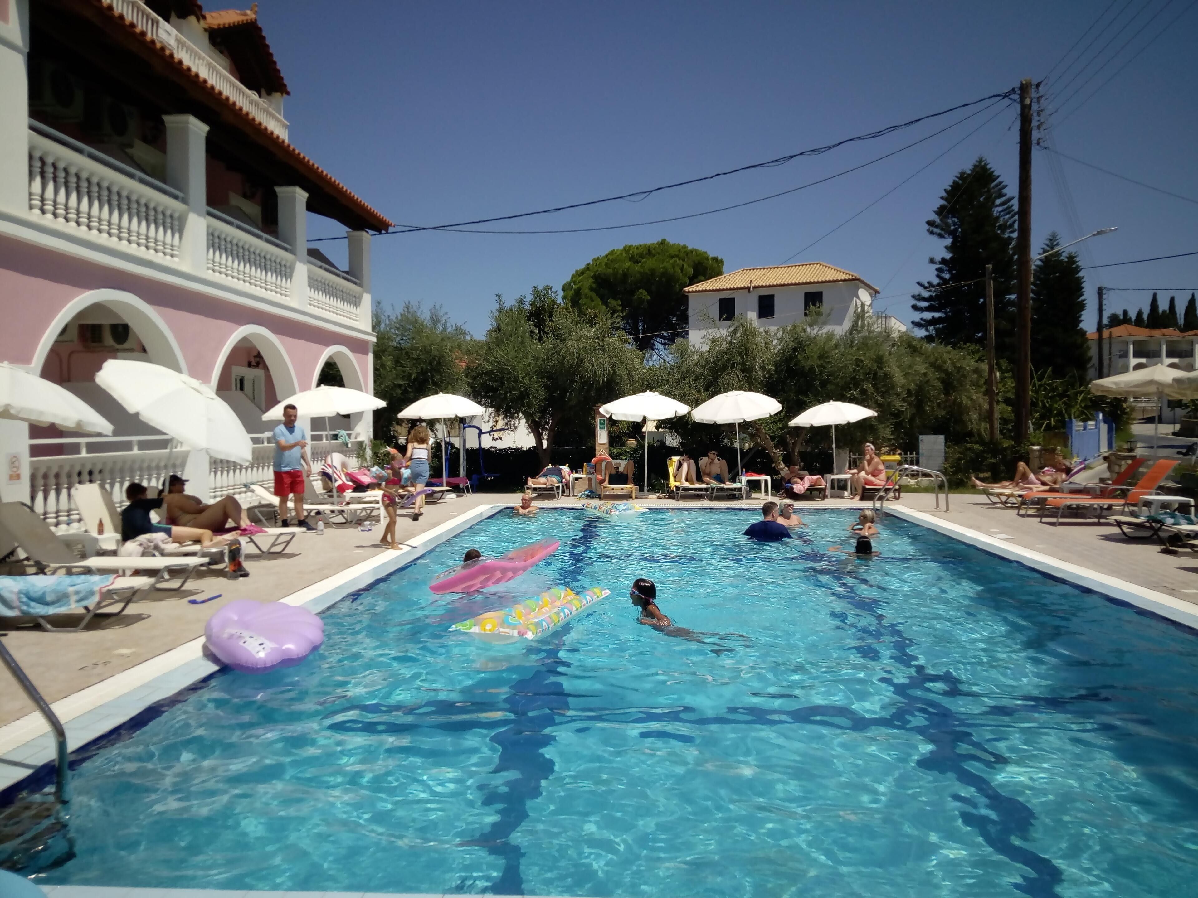 Une piscine où des gens nagent et se prélassent. Des parasols et des chaises longues sont disposés autour de la piscine.