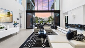 A modern living room with a large sectional sofa, a coffee table, and a large window offering a view of the cityscape. - Boardwalk Mansion Lauderdale Villa: Private Pool, Hot Tub, Mini-Golf & Guest House (Fort Lauderdale)
