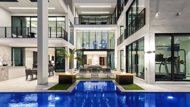 A modern indoor swimming pool with a central seating area, surrounded by glass walls and multiple levels of balconies. - Boardwalk Mansion Lauderdale Villa: Private Pool, Hot Tub, Mini-Golf & Guest House (Fort Lauderdale)