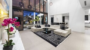 A modern living room with a sectional sofa, a coffee table, and a black and white rug. There is a large window with a view of palm trees and a pool. - Boardwalk Mansion Lauderdale Villa: Private Pool, Hot Tub, Mini-Golf & Guest House (Fort Lauderdale)
