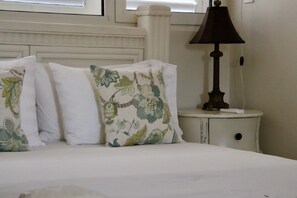 A bed with white bedding and floral-patterned pillows, a bedside table with a lamp, and a window with curtains.