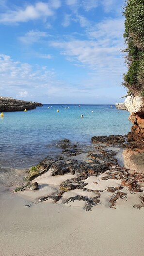 A coastal beach with clear blue water, rocky shoreline, and a cliff with vegetation.