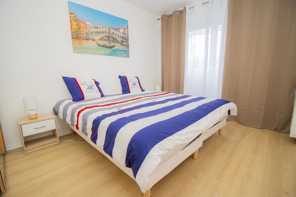 Uma cama bem arrumada com colcha listrada, uma mesa de cabeceira com abajur e um quadro de uma ponte na parede.