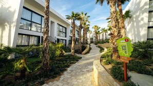 A pathway lined with palm trees leading to a modern building with large windows and a sign indicating exit and village locations. - Alannia Salou Resort  (Salou)