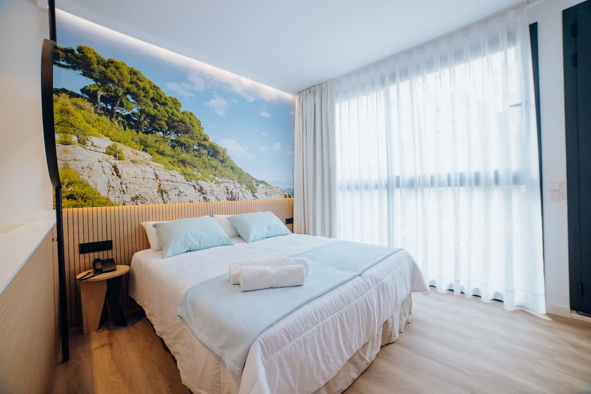 A modern bedroom with a large bed, a nightstand, a wall-mounted phone, and a view of a rocky landscape through sheer curtains.