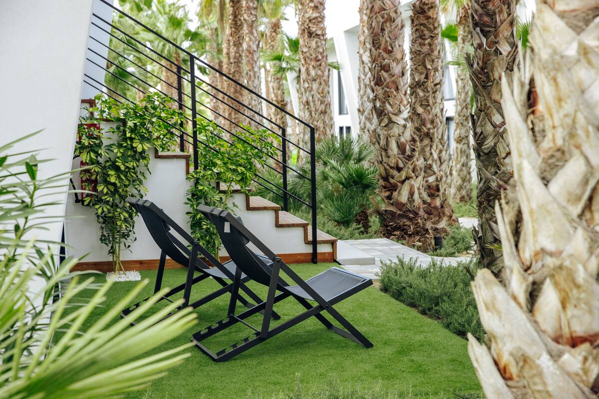 A modern outdoor area with a black lounge chair, a staircase, and lush greenery including palm trees and a white wall with climbing plants.