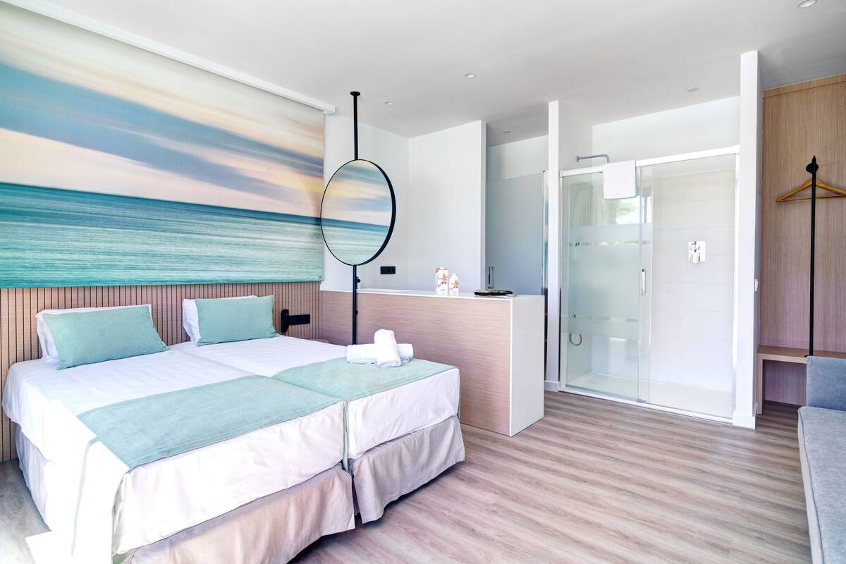 A modern hotel room with a large bed, a desk, a glass shower area, and a view of the sea.