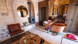 Una habitación rústica con paredes de piedra, una bañera de cobre grande, un sillón de cuero y una mesa redonda de madera con varios objetos.
