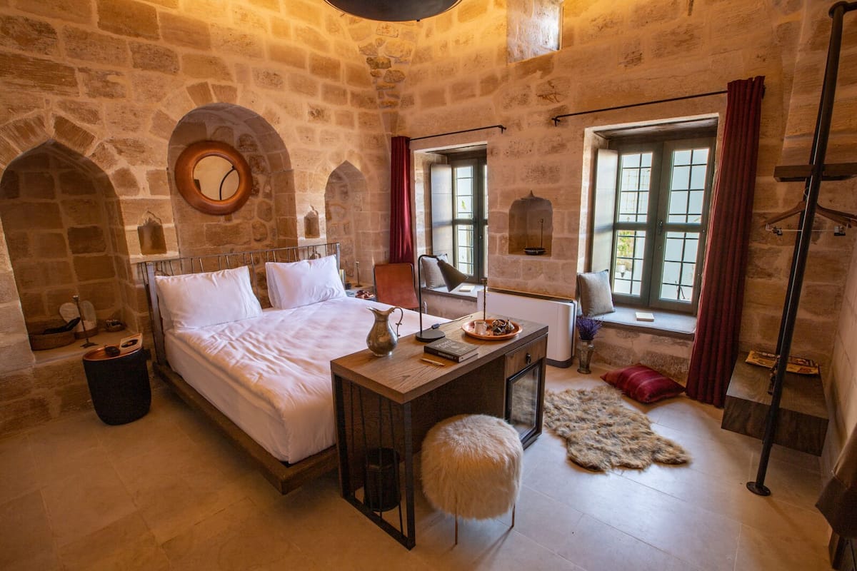 A stone-walled bedroom with a bed, a small table, a chair, and a window with curtains.