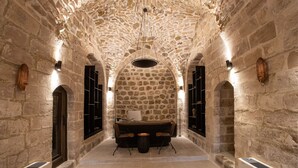 Un pasillo con paredes de piedra, una mesa y sillas, arcos y lámparas fijadas a la pared.